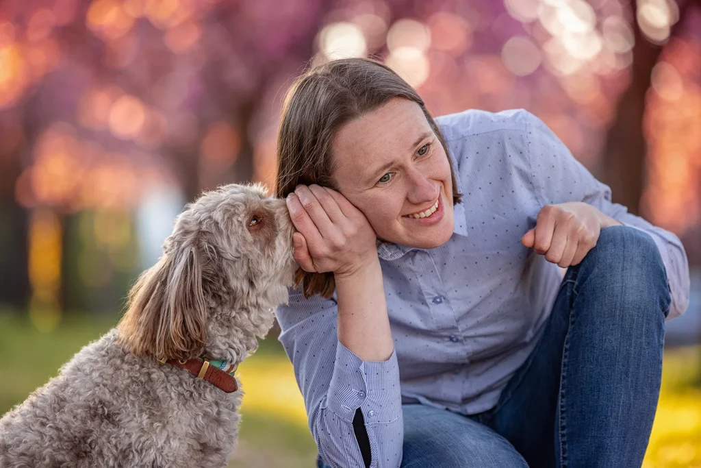 Ein Mensch und ihr Aussiedoodle sitzen in Emsdessten bei Münster (NRW) im Gras unter den blühenden Kirschbäumen. Der Hund scheint der Halterin etwas ins Ohr zu flüstern. Fotografiert von der Hundefotografin Anne Bertling aus Ascheberg im Münsterland.