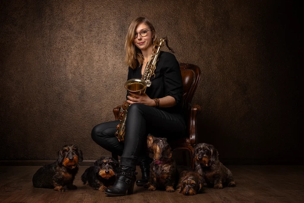 EIne Hundehalterin sitzt mit ihrem Saxophon und ihren fünf Dackeln auf einem Ledersessel vor einem barocken Hintergrund. Der Boden ist aus Holz, das Bild wirkt wie aus der Renaissance, fotografiert im Fotostudio in Ascheberg bei Münster, NRW, von der Hundefotografin Anne Bertling.